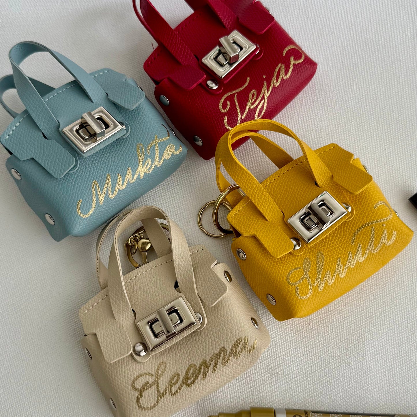 Four small handbags in different colors with names written on them, on a white surface.