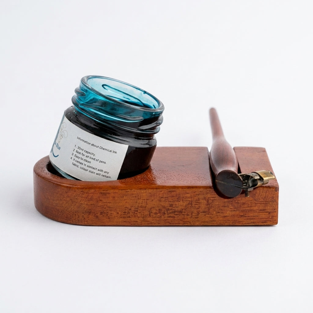A dark glass jar of ink resting on a wooden ink stand with a wooden stylus and brass clip, photographed on a clean white background.
