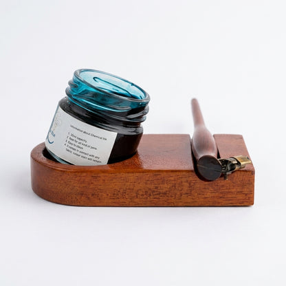 A dark glass jar of ink resting on a wooden ink stand with a wooden stylus and brass clip, photographed on a clean white background.