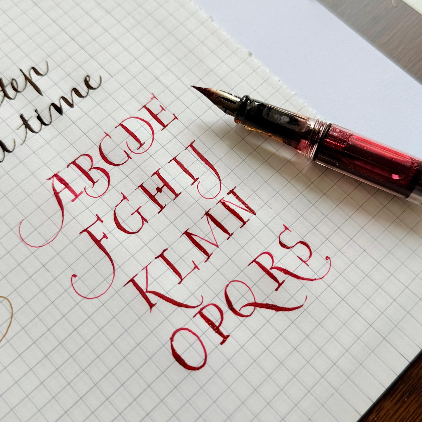 Calligraphy practice sheet with red ink letters and a pen on grid paper.