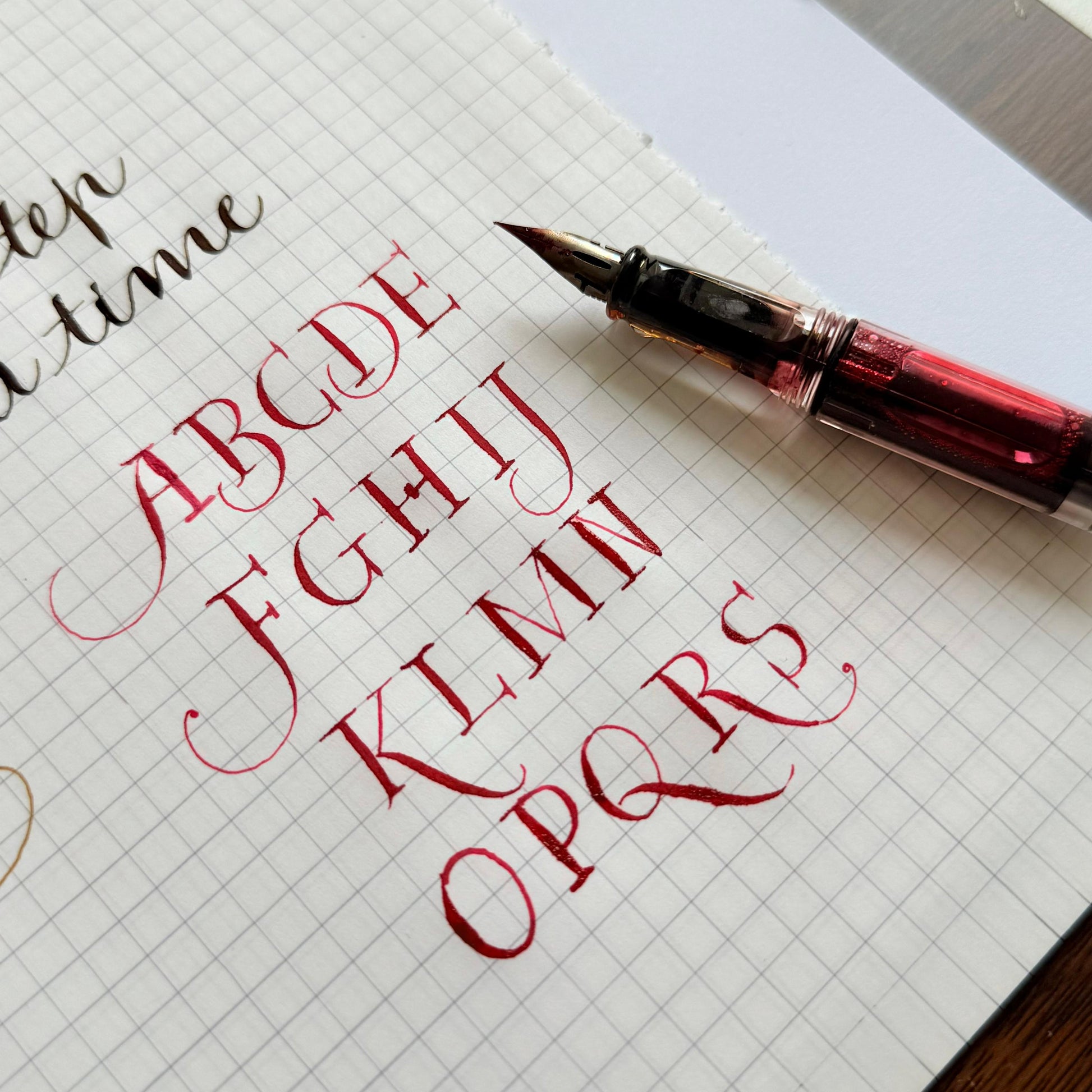 Calligraphy practice sheet with red ink letters and a pen on grid paper.