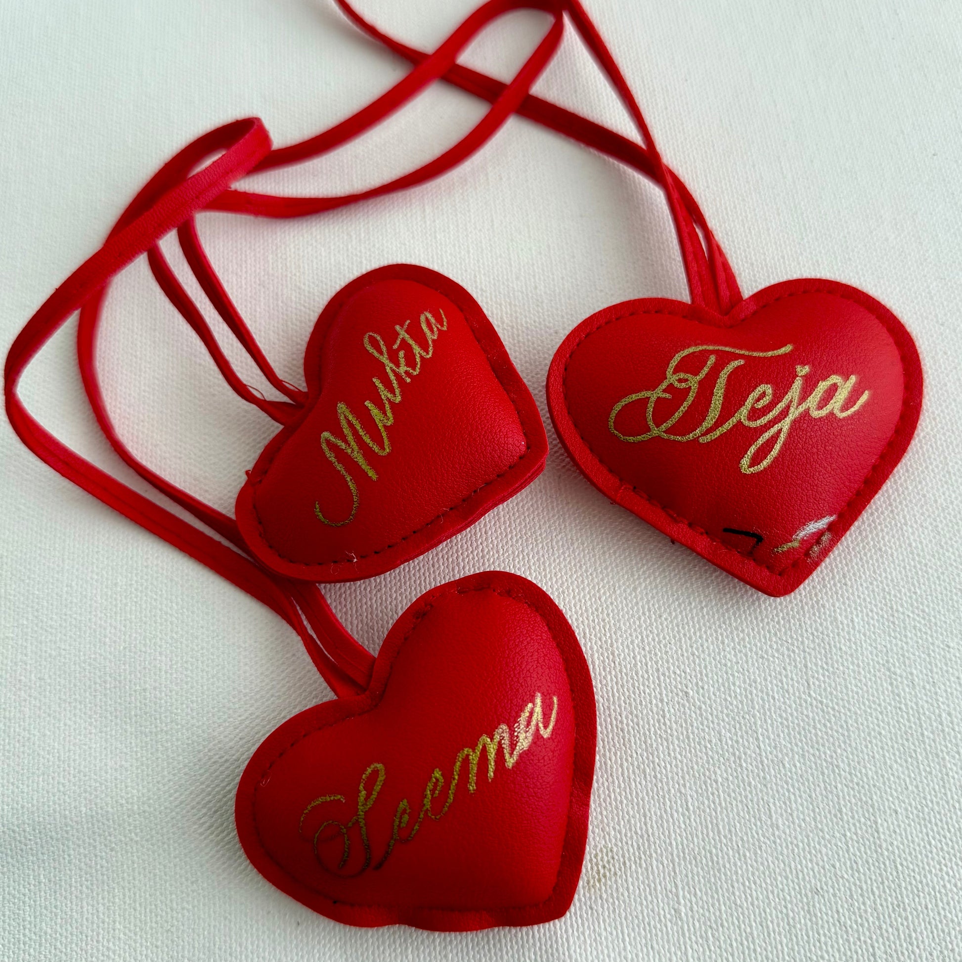 Three red heart-shaped ornaments with names embroidered on them, on a white background.